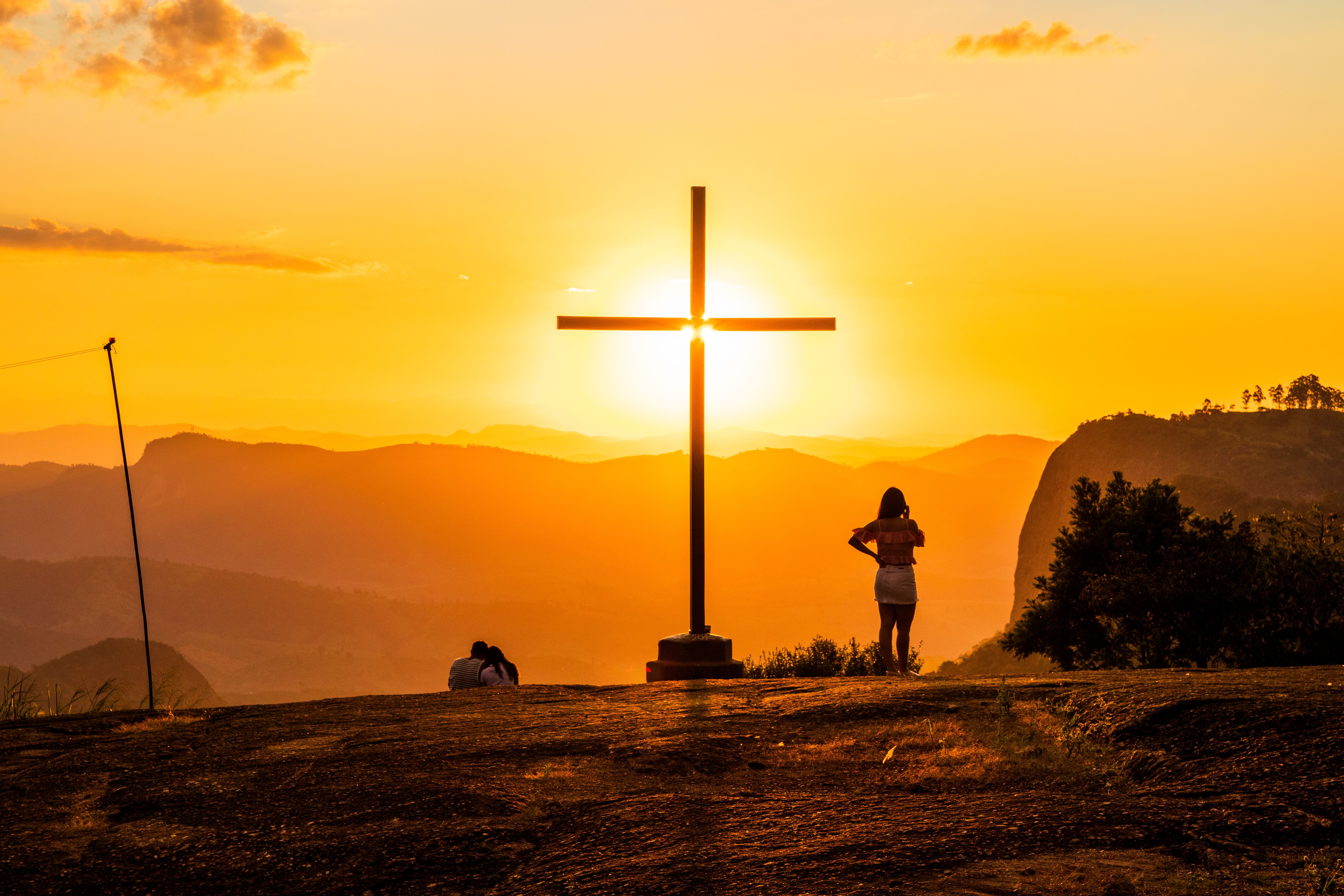 Back view of anonymous travelers standing near cross behind mountains under colorful sky with shining sun at sunset — Foto: Marcos Luis Leonardo / Pexels