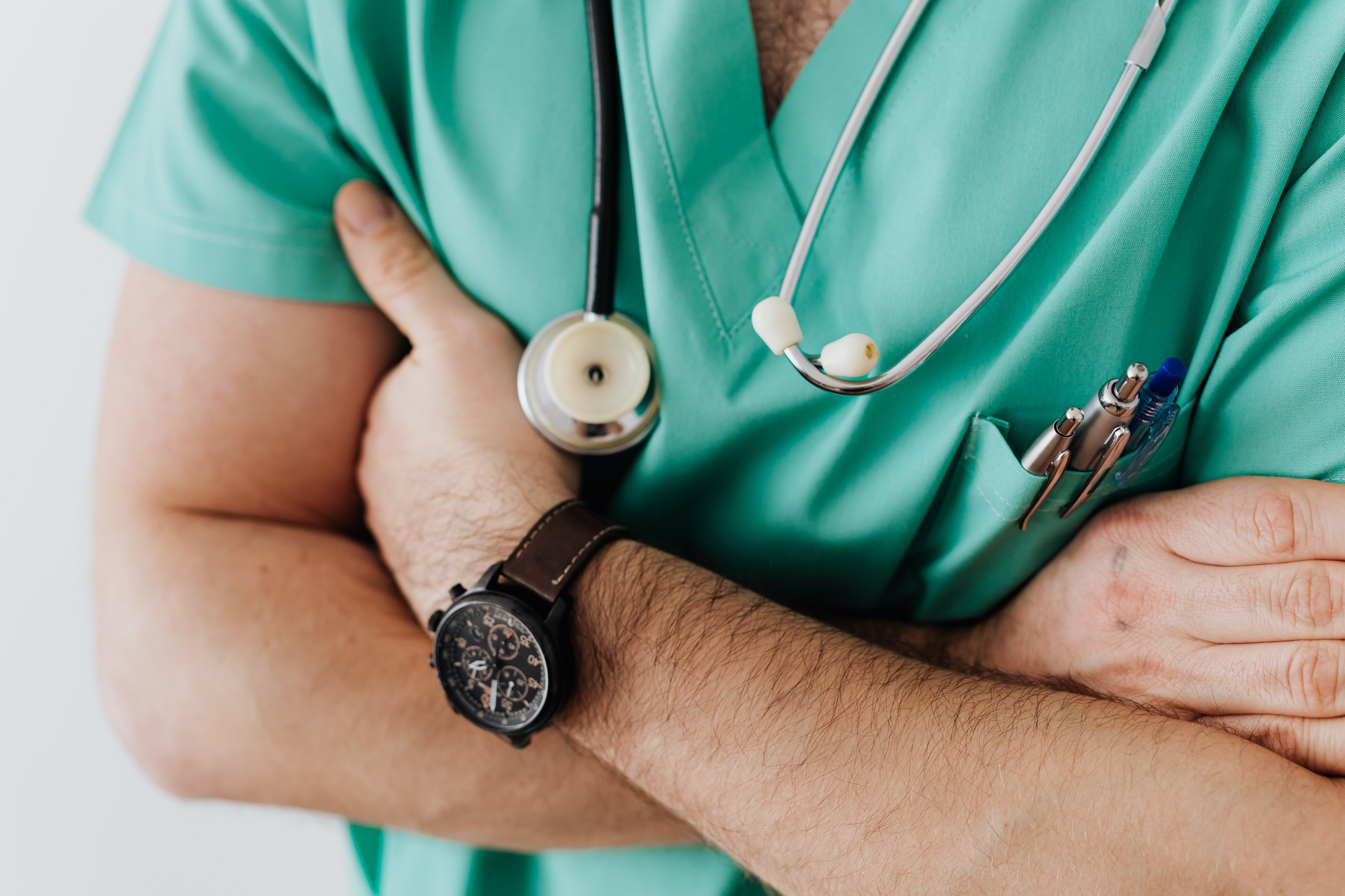 Unrecognizable man wearing medical uniform with stethoscope and pens standing in hospital with crossed arms during work in day time — Foto: https://kaboompics.com/ / Pexels